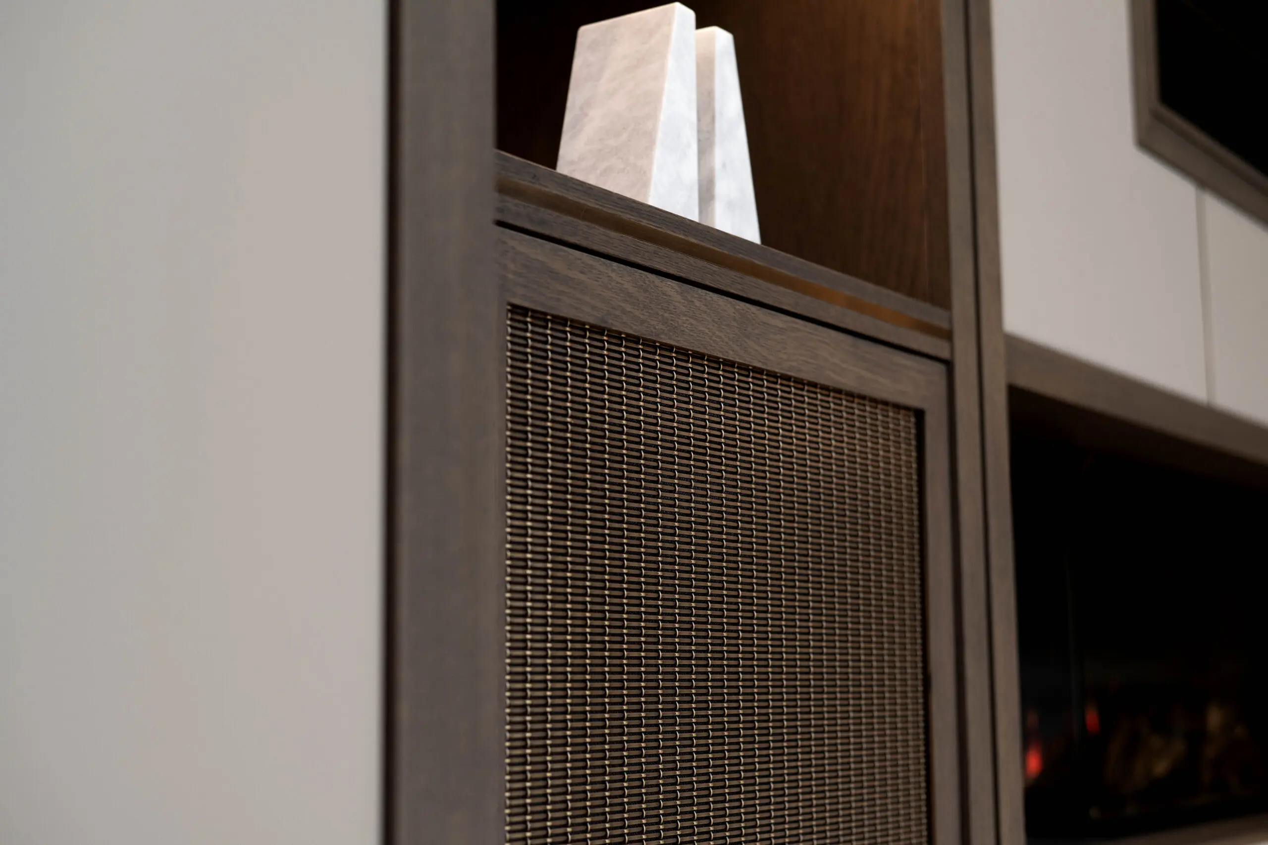 Close-up of a modern cabinet with dark wood accents. A Gauze- textured metal panel is prominent, while two white, pyramid-shaped objects sit on the shelf above.