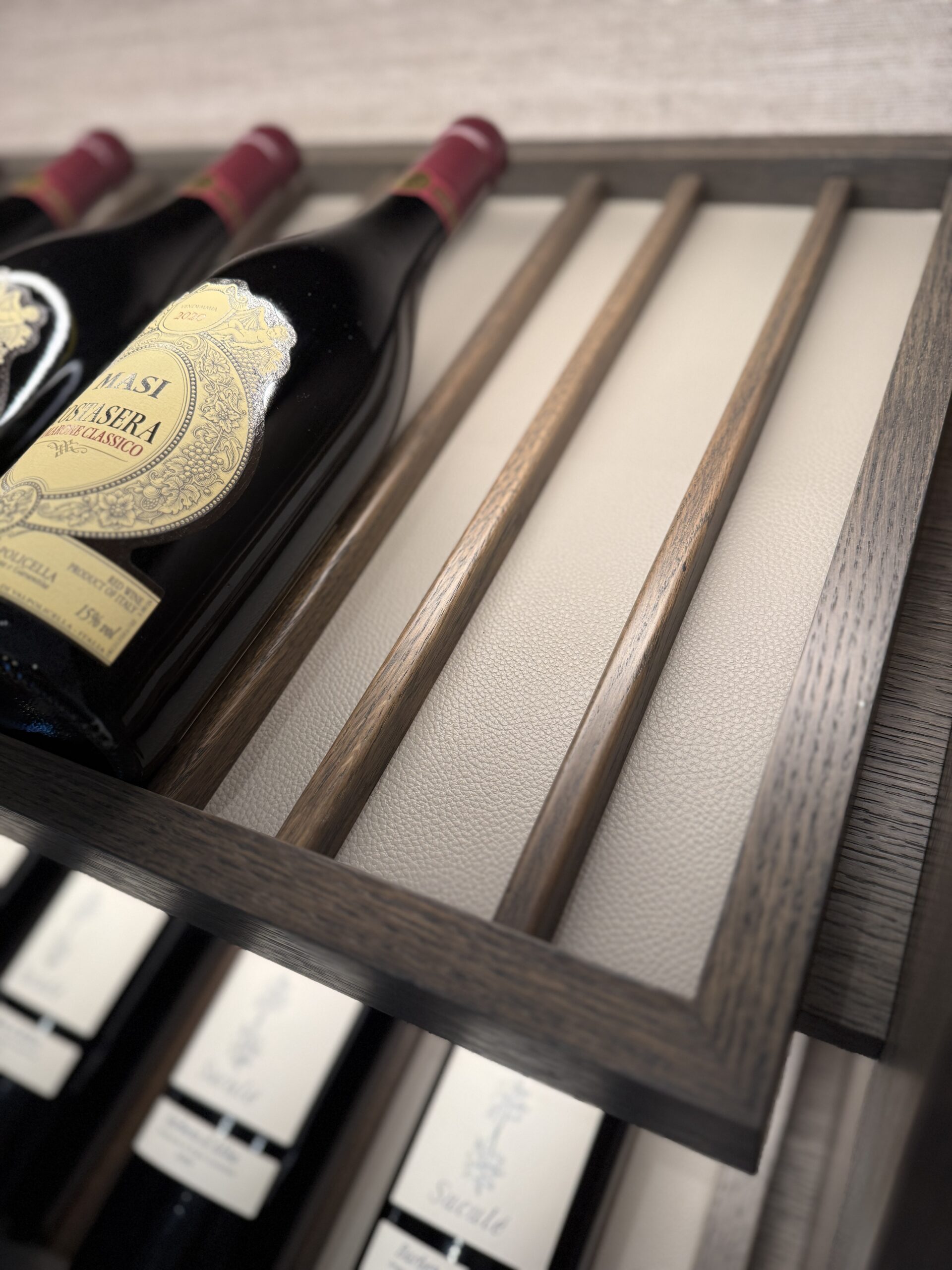 Close-up of wine bottles with ornate labels displayed on a wooden rack lined in a light grey leather. The perspective shows textured wood and a leather-like backdrop, evoking elegance.