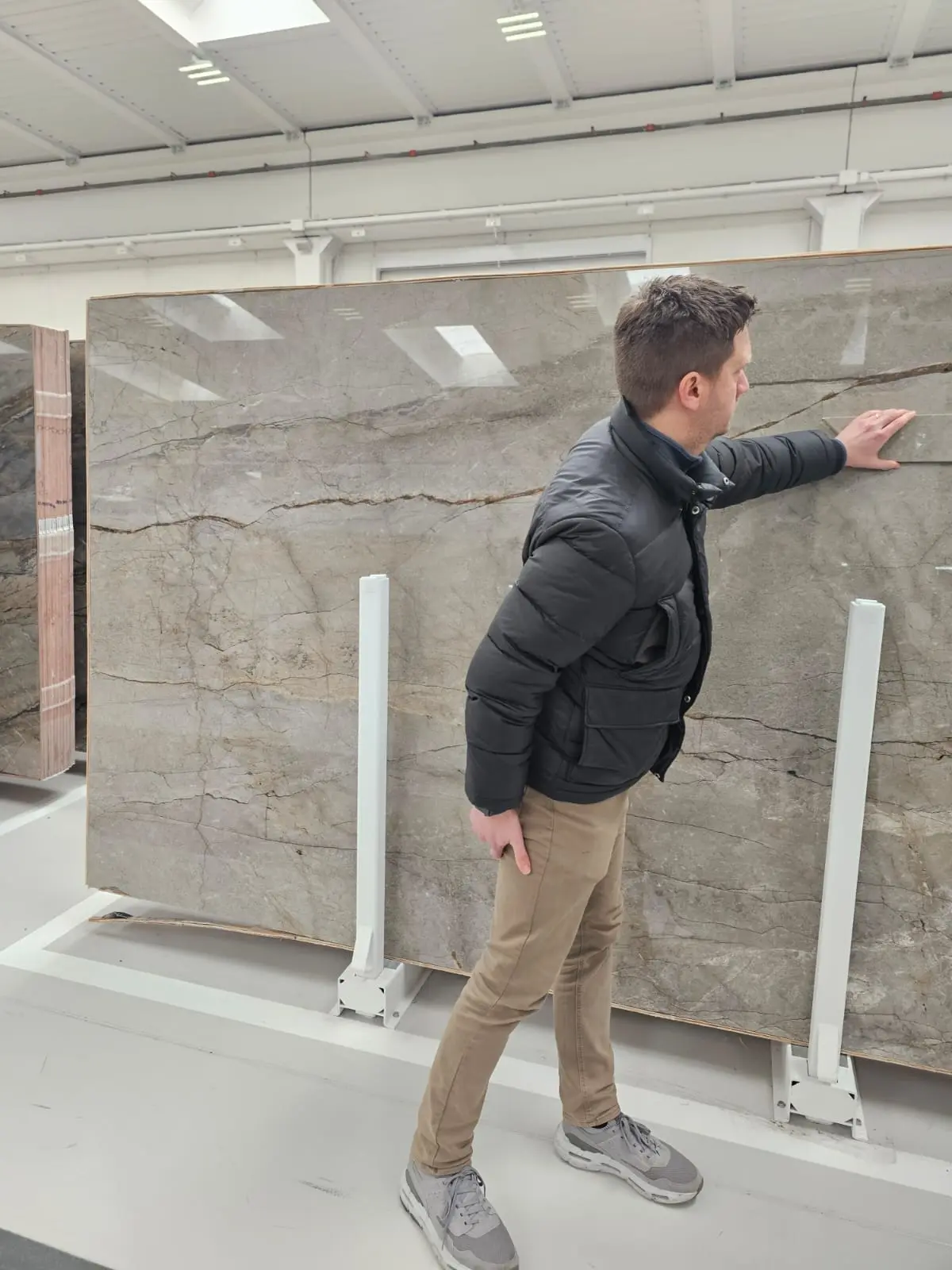 Niall, the lead Project Designer, examines a large slab of marble with grey and brown veins in a brightly lit warehouse in Verona, conveying curiosity.
