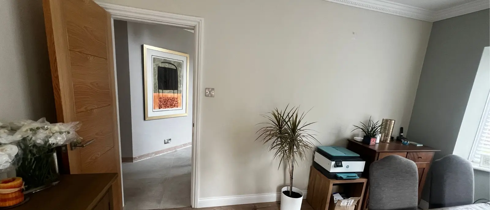 Before; A dark office room with a wooden desk, chairs, and a printer. A potted plant adds greenery. Open door leads to hallway with framed art on the wall.