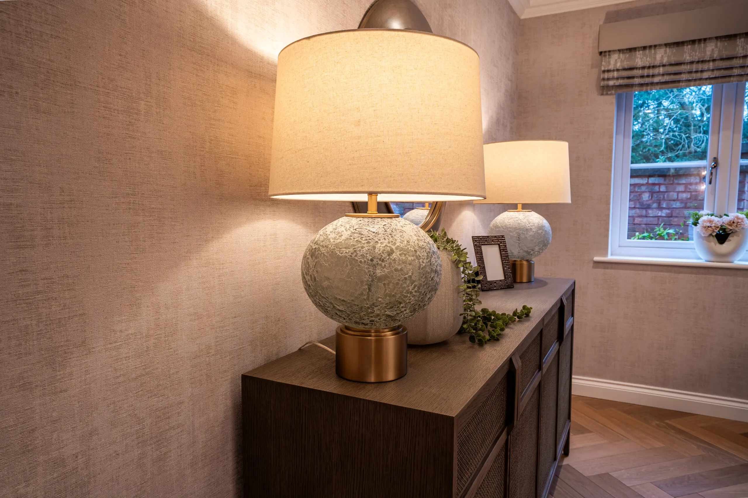 Elegant room with textured wallpaper and warm lighting. A spherical lamp with a gold base sits on a wooden sideboard, complemented by a framed mirror and window.