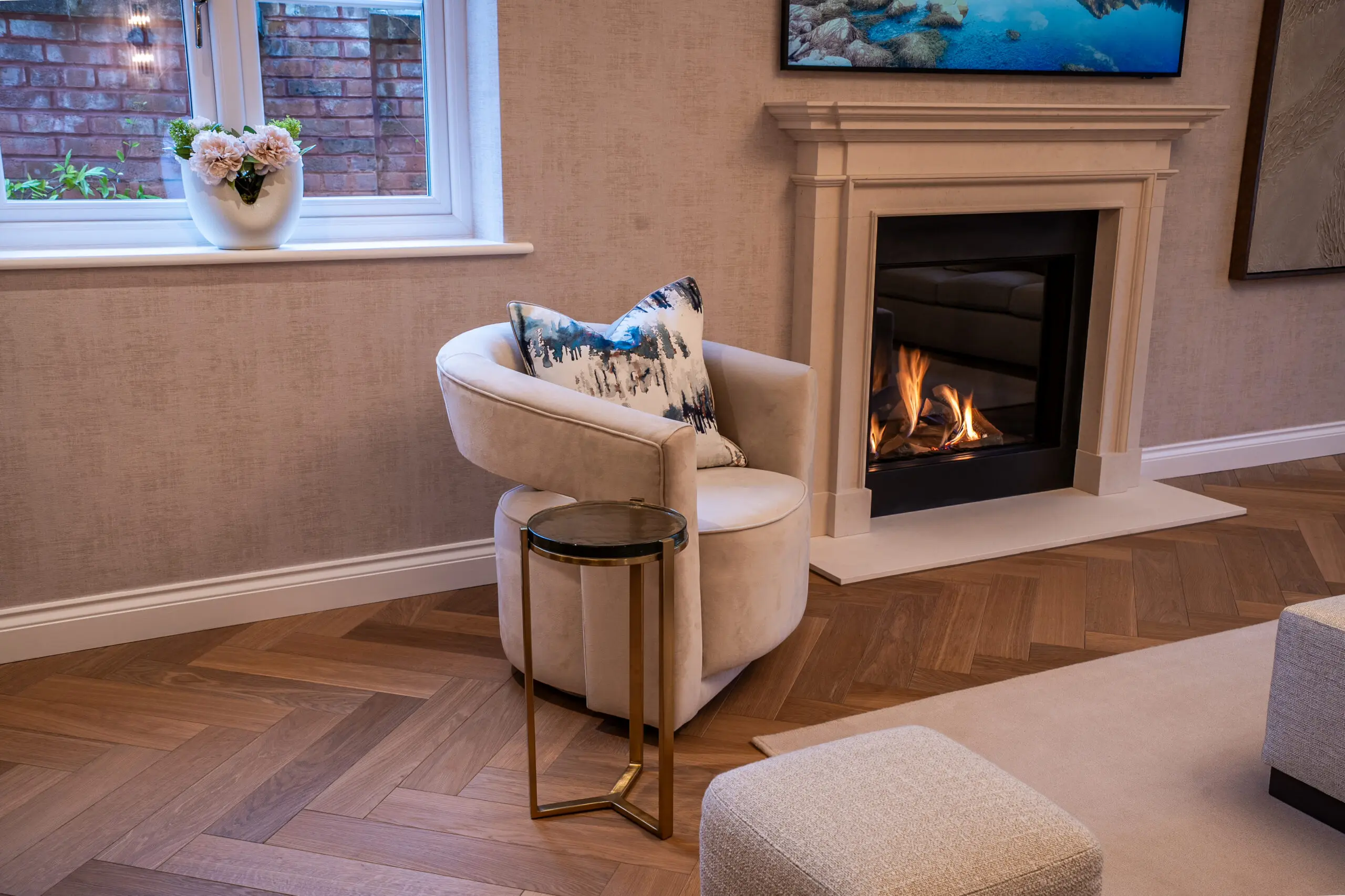 A cozy living room with a beige armchair and cushion beside a lit fireplace. A small round table is nearby, and a window with flowers adds a warm touch.