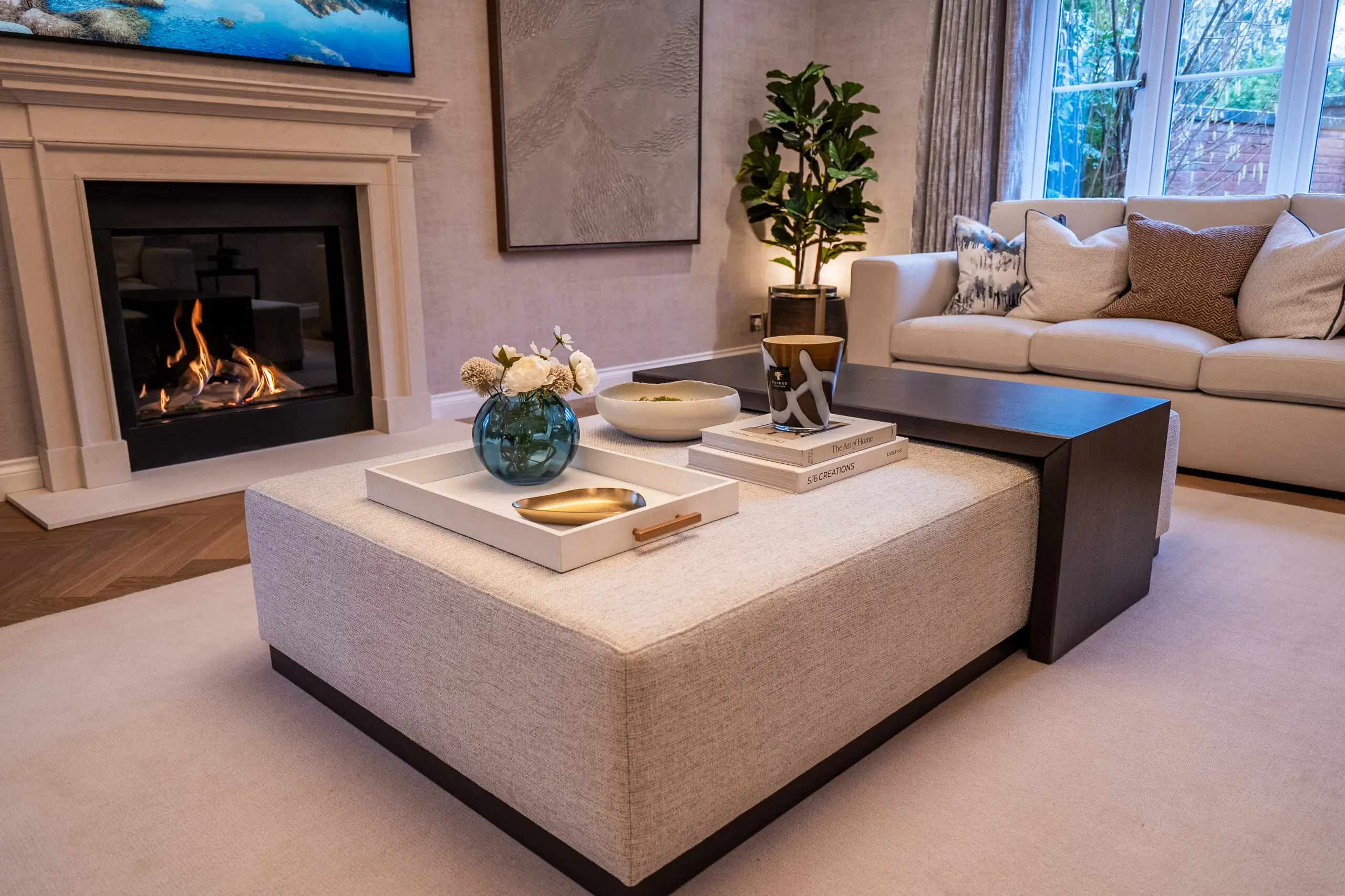 Elegant living room with a cozy fireplace, plush beige sofa, large ottoman with decor, and a green plant. Warm and inviting ambiance.