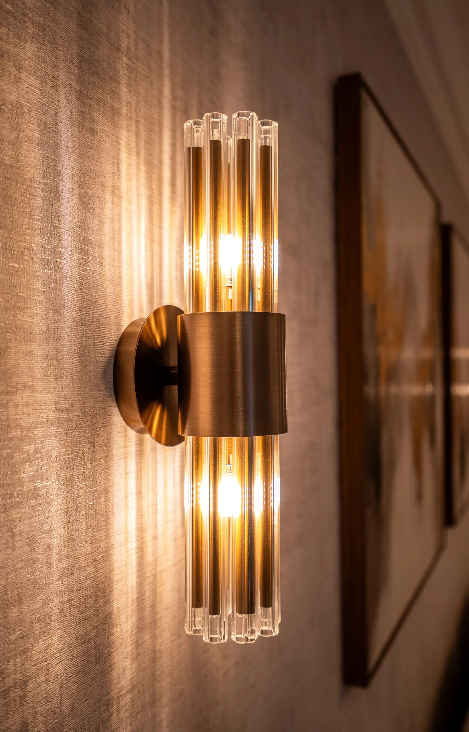 Art Deco wall sconce with cylindrical glass tubes emits warm light on a textured beige wall, enhancing a cozy and elegant interior atmosphere.