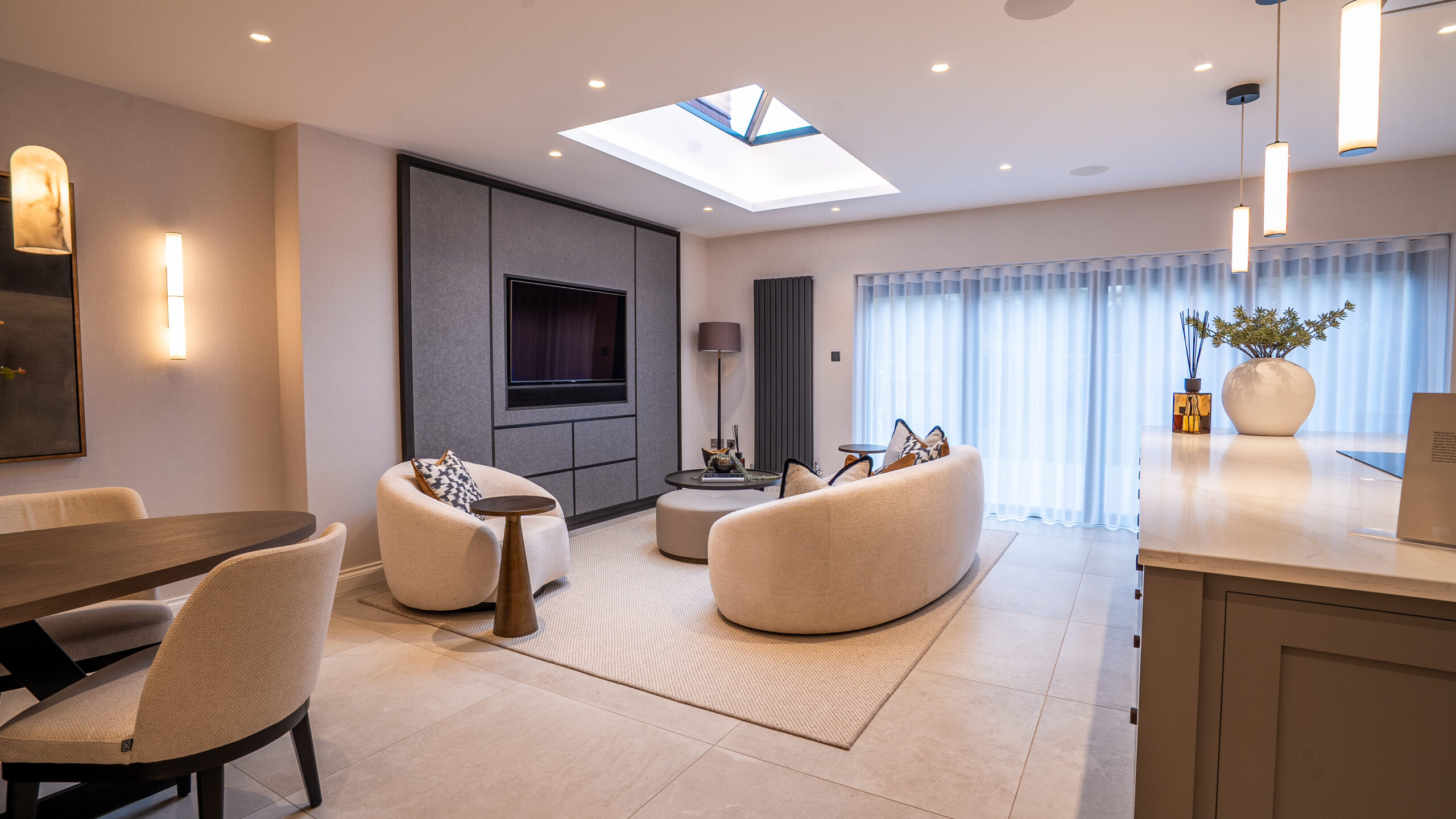 Avonrise ground floor renovation: Modern living room with neutral tones, curved sofas, a wall-mounted TV, skylight, dining area, large windows with sheer curtains, and contemporary lighting fixtures. 721