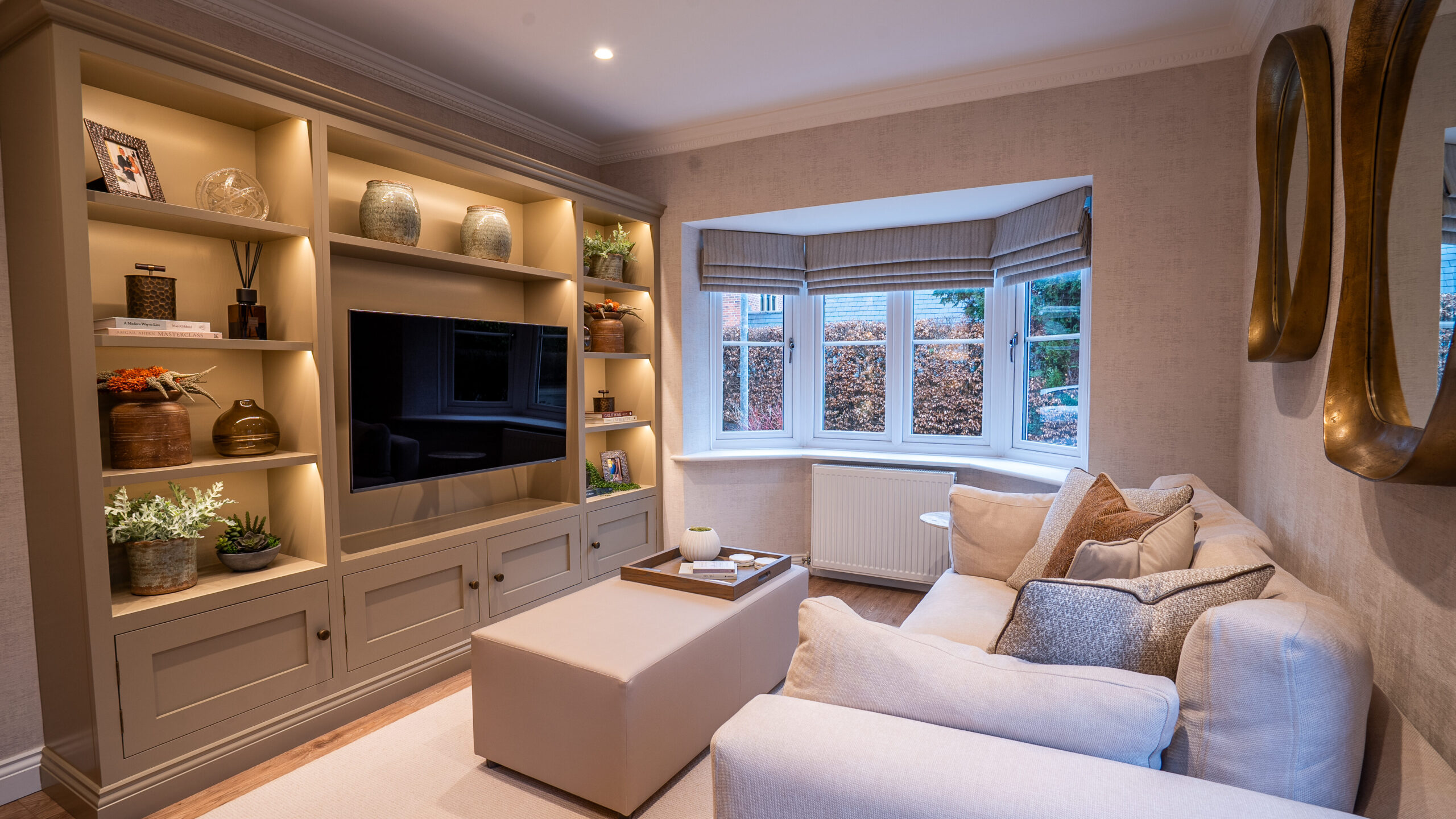 Elegant living room with cozy beige sofas and a large ottoman. Built-in shelves with decor items flank a TV. The bay window adds natural light