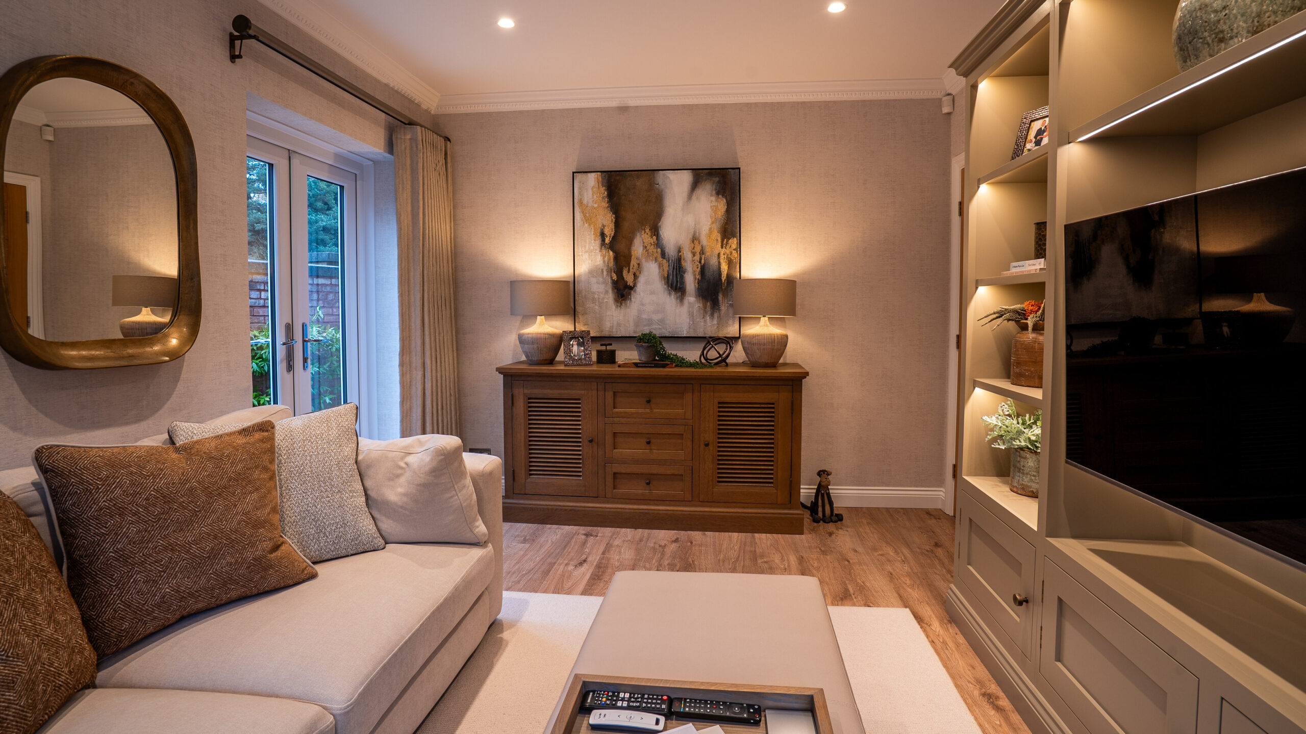 A cozy living room with a light beige sofa adorned with textured cushions, beside glass doors. A painting above a wooden sideboard adds elegance. A built-in shelf with decor items enhances the warm ambiance.