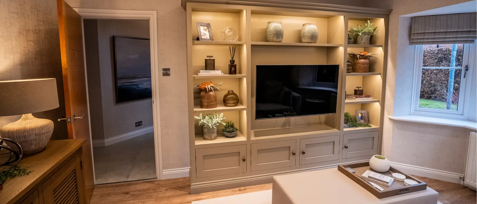 After: Modern living room with neutral tones, featuring a built-in shelving unit displaying decorative items, a flat-screen TV, and a soft-lit ambiance.