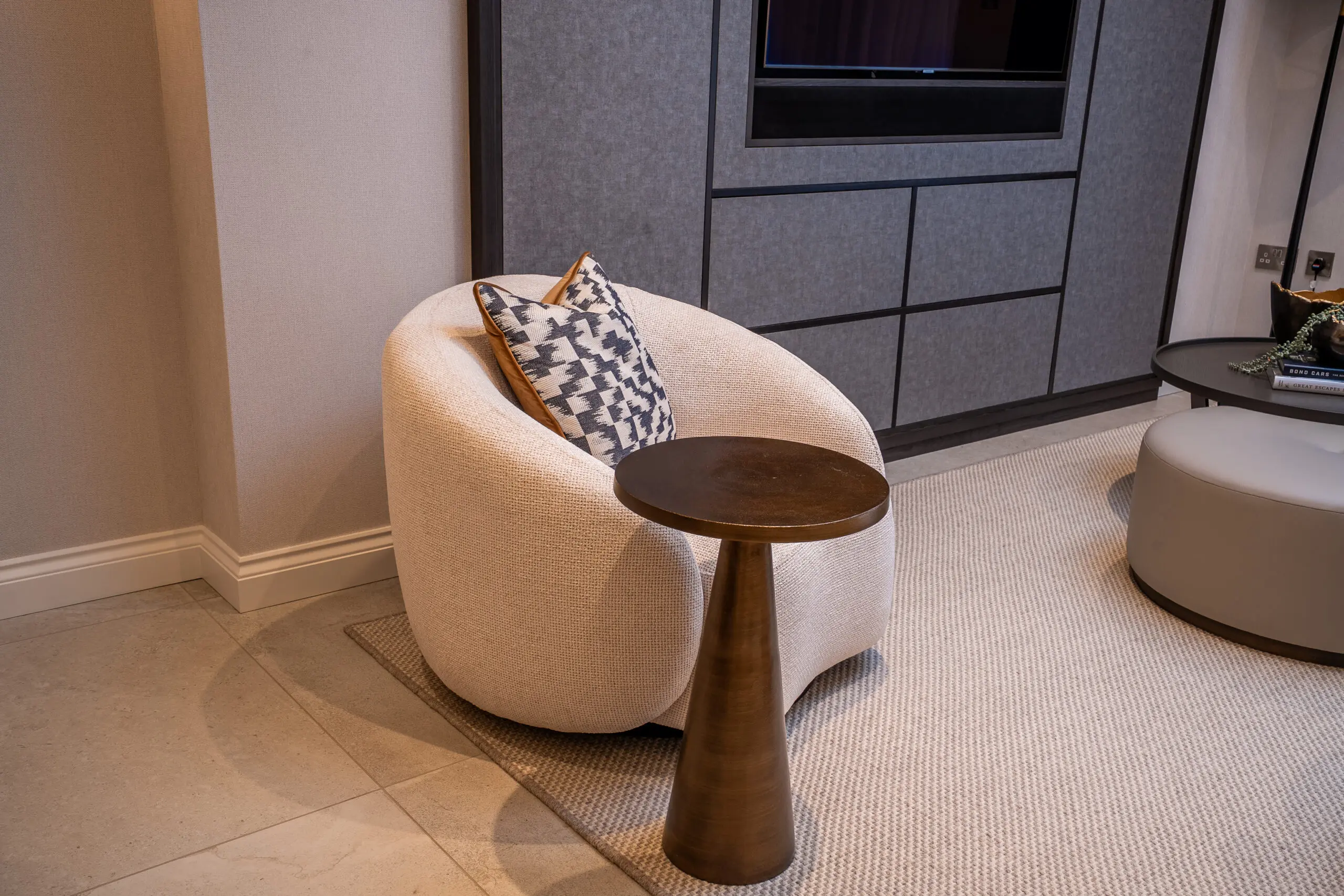 A cozy, modern room with a round, textured white chair, resting a patterned cushion. A small bronze side table stands beside, adding elegance. Comfortable ambiance.