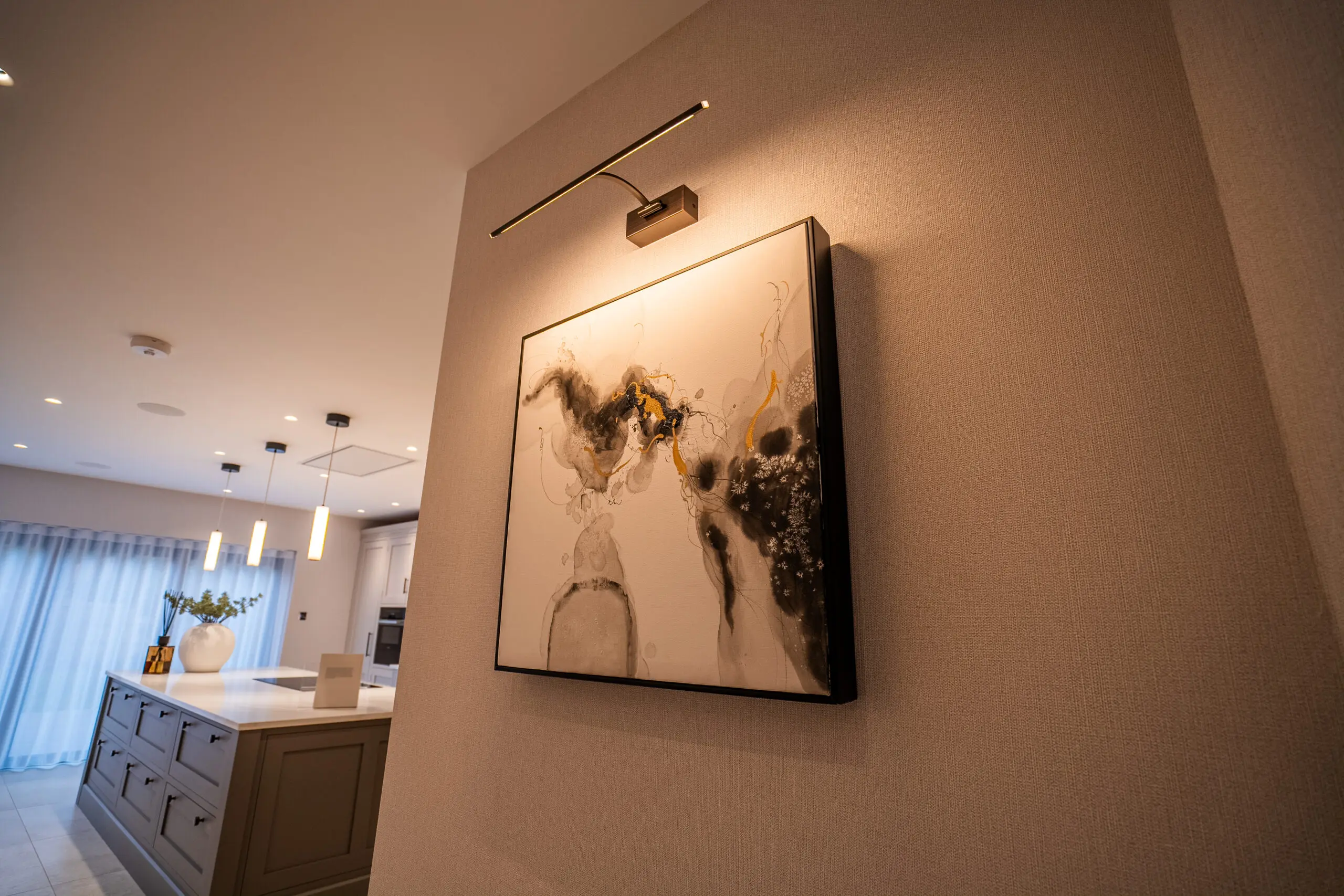 Modern kitchen with abstract art illuminated by a wall light. Features sleek cabinets, pendant lights, and a vase on the island, creating a cozy atmosphere.
