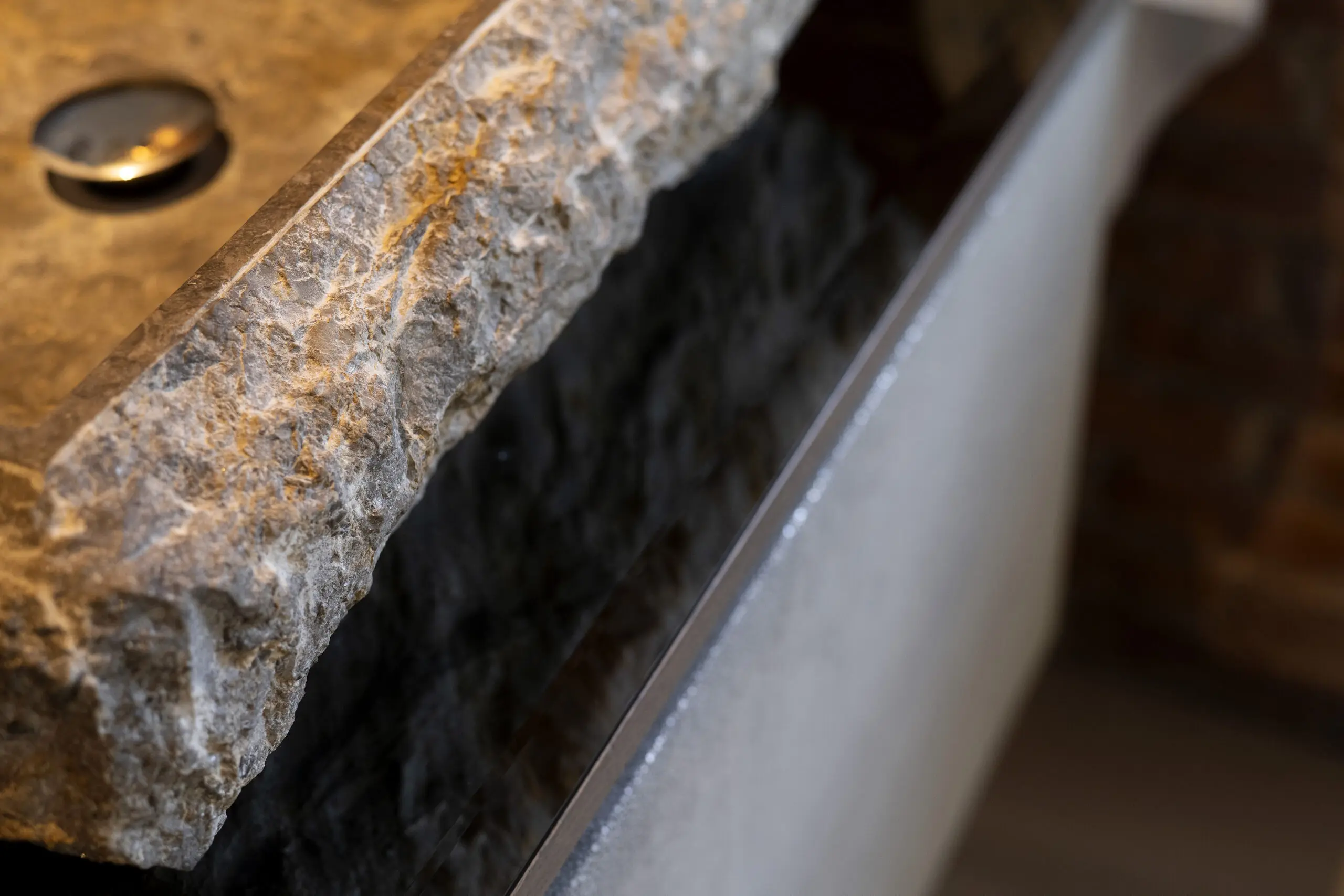 Close-up of a textured stone sink with a rough, natural edge as water flows over the surface. A round drain is visible on the left, and the smooth basin contrasts with the rugged stone detail.