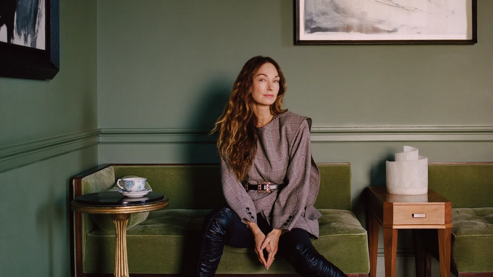 A woman with long wavy hair sits on a green upholstered bench against a green wall, wearing a patterned top and dark pants. A small round table with a teacup is on the left, and a wooden side table with a sculptural object is on the right. Framed artwork hangs above. Source: Home & Gardens.