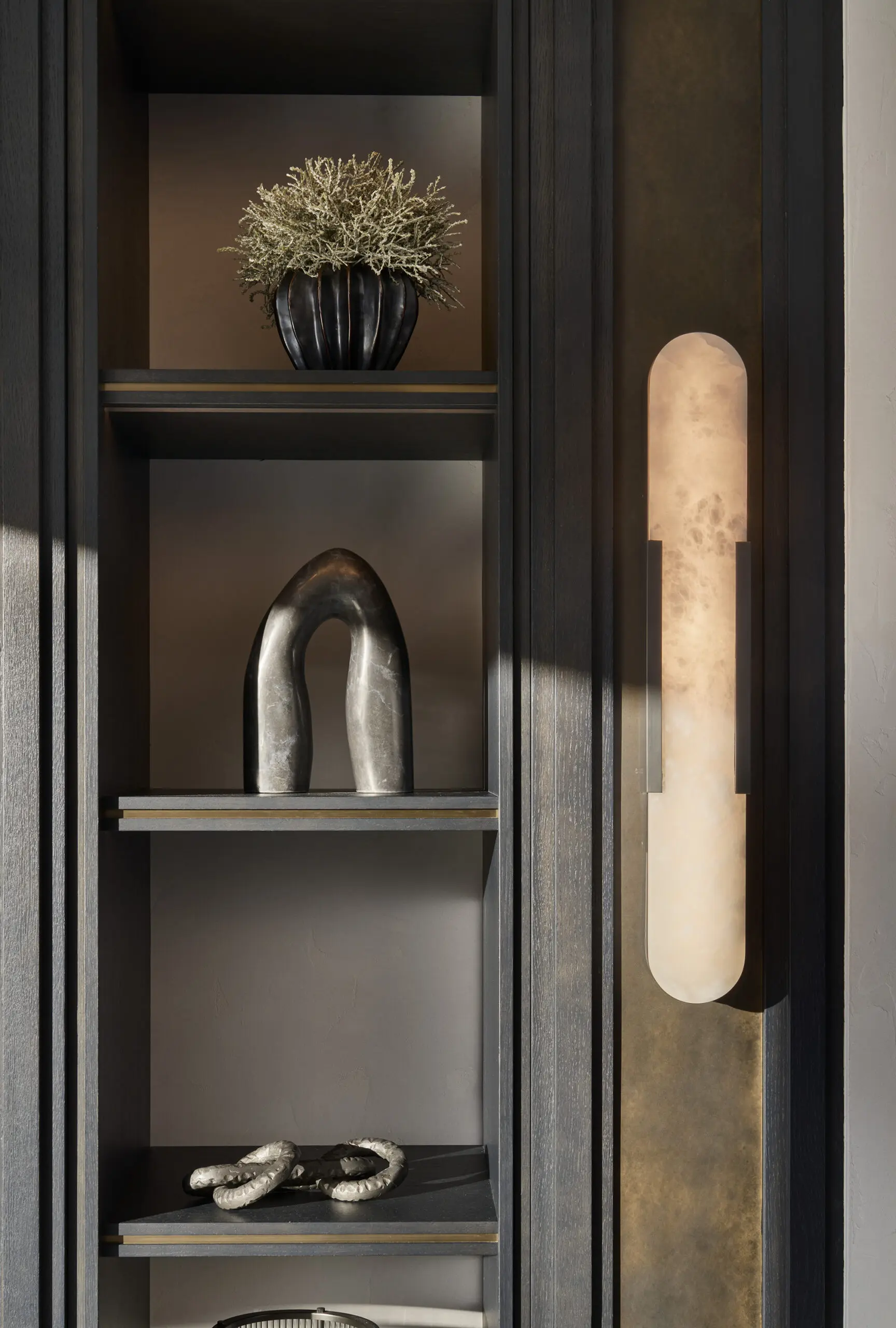 A dark, modern shelf holds a potted plant, abstract metal sculptures, and a soft-glowing vertical wall light, creating a serene and elegant ambience.