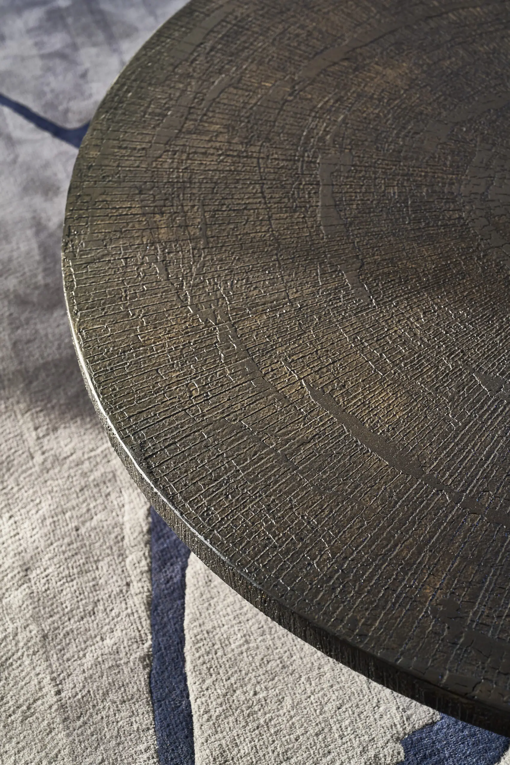 Close-up of a round wooden table with a textured, cracked surface. The tabletop rests on a light gray fabric, creating a rustic, natural atmosphere.