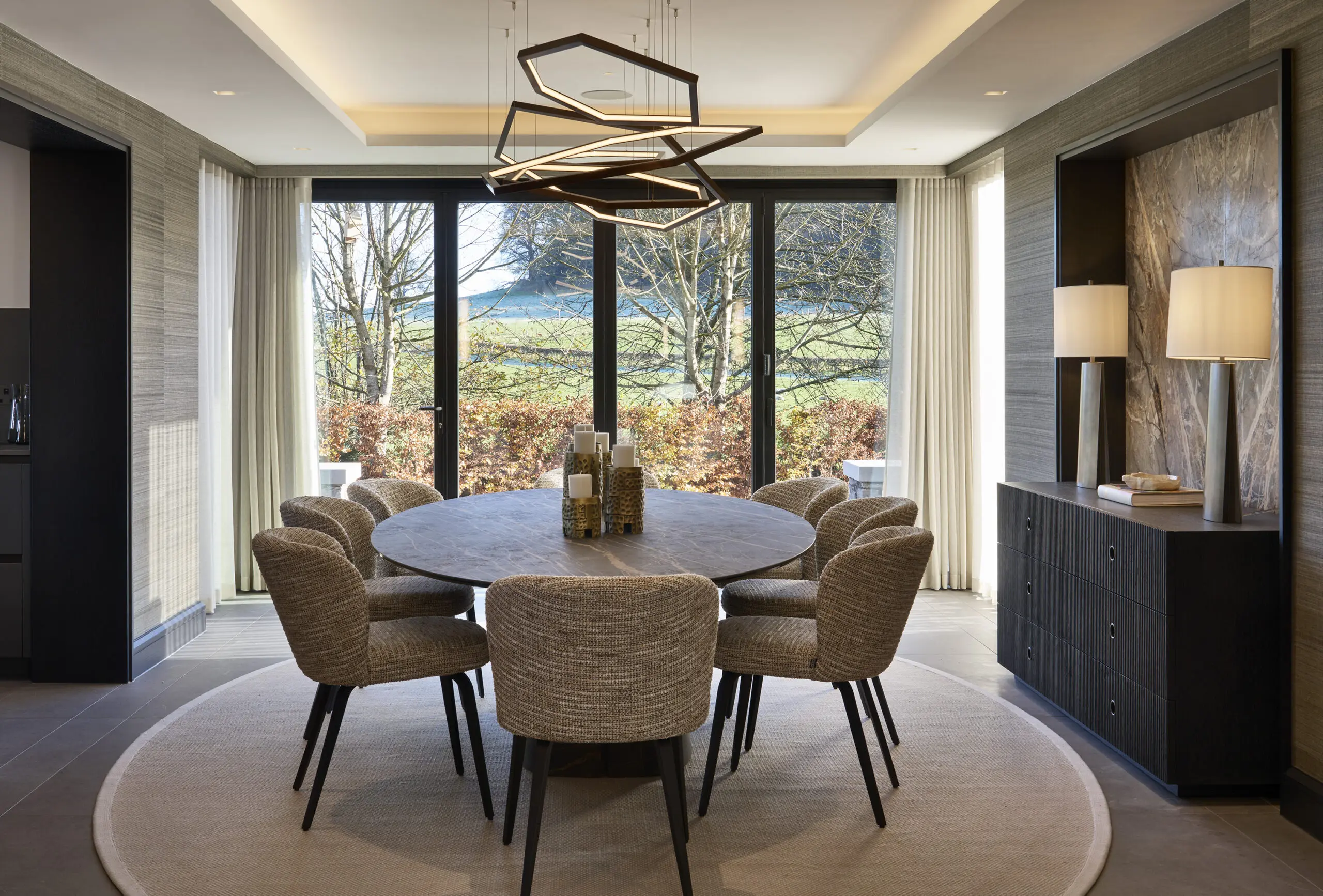 Modern dining room with a round table and eight textured chairs, lit by a geometric chandelier. Large windows reveal a serene outdoor view.