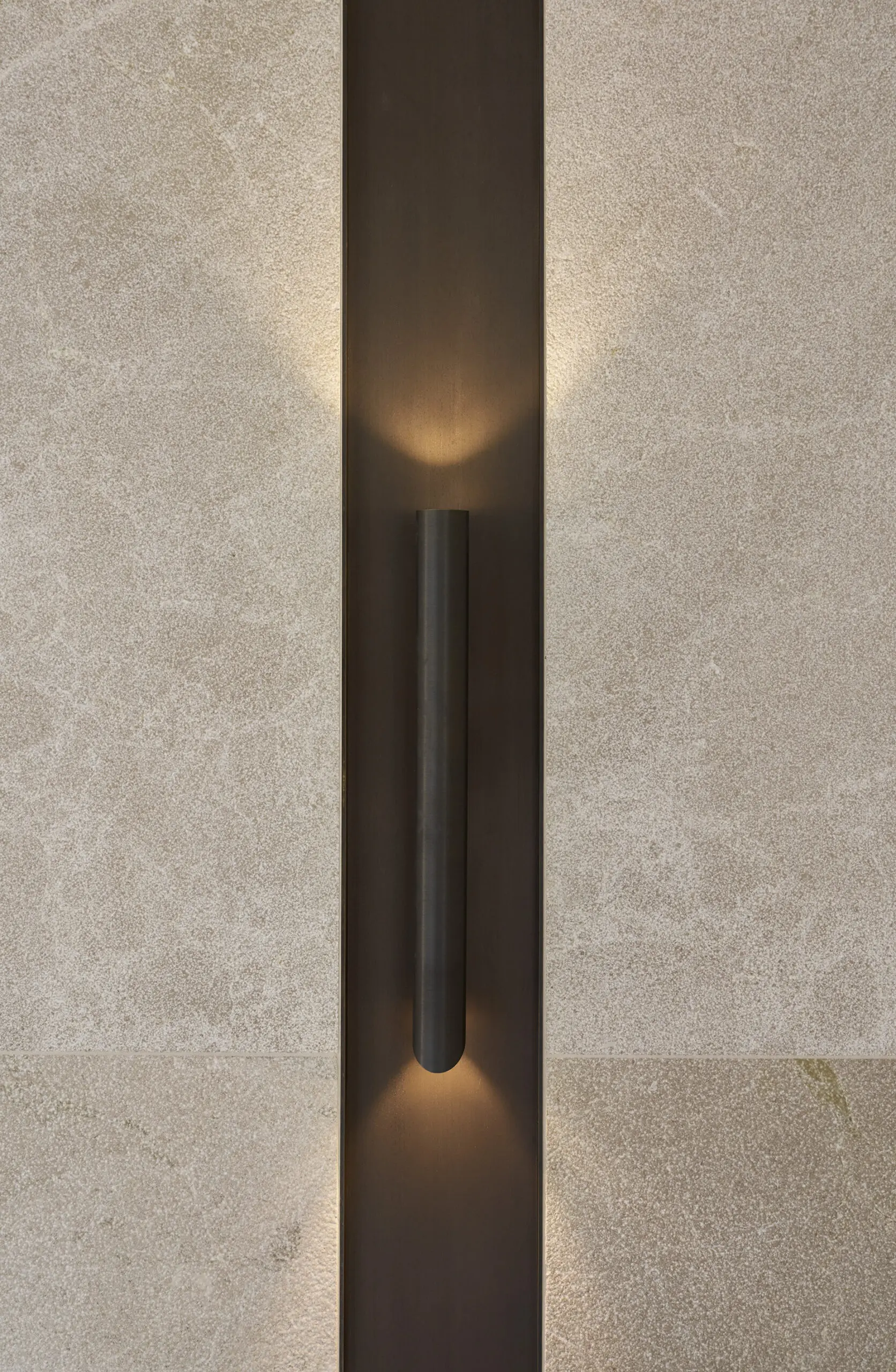 A sleek black wall sconce emits warm light, creating a cozy ambiance on a light, textured beige wall. The minimalist design adds modern elegance.