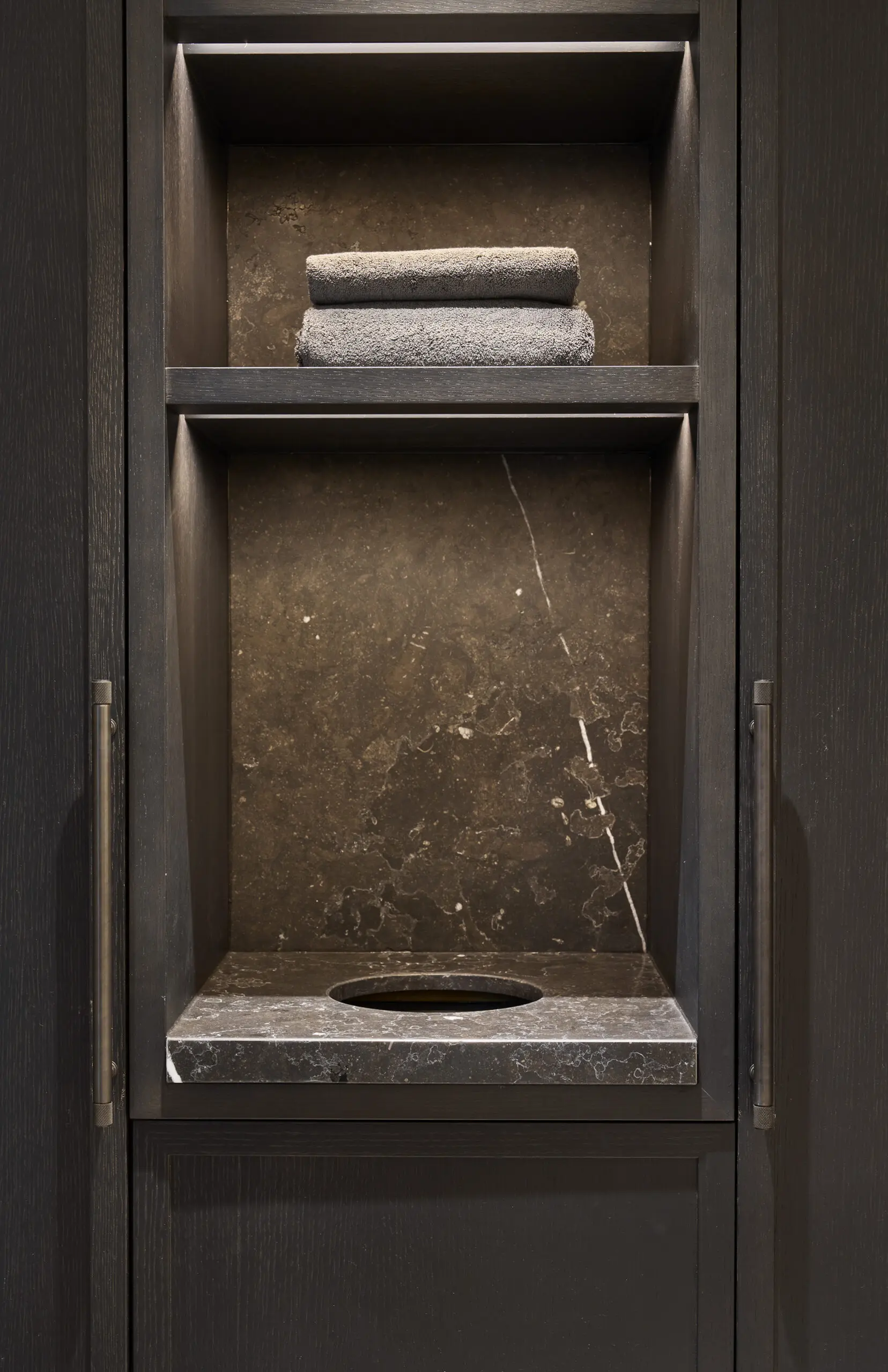 Sleek dark-colored shelving unit with two folded gray towels on the top shelf and a circular cutout in the marble-textured surface below.