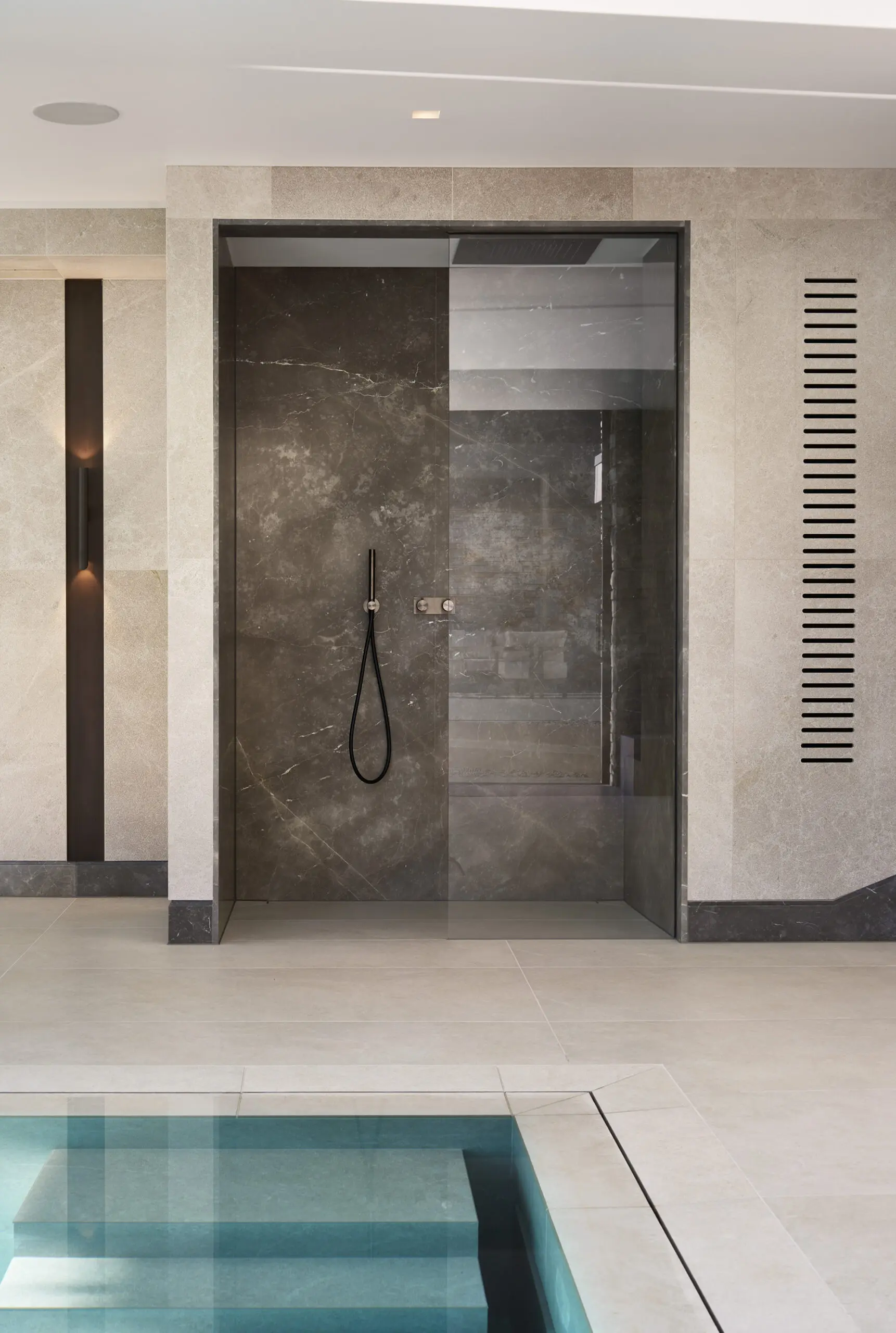 Luxurious spa room featuring a glass-enclosed shower with sleek black fixtures, surrounded by marble tiles, adjacent to a serene turquoise plunge pool.