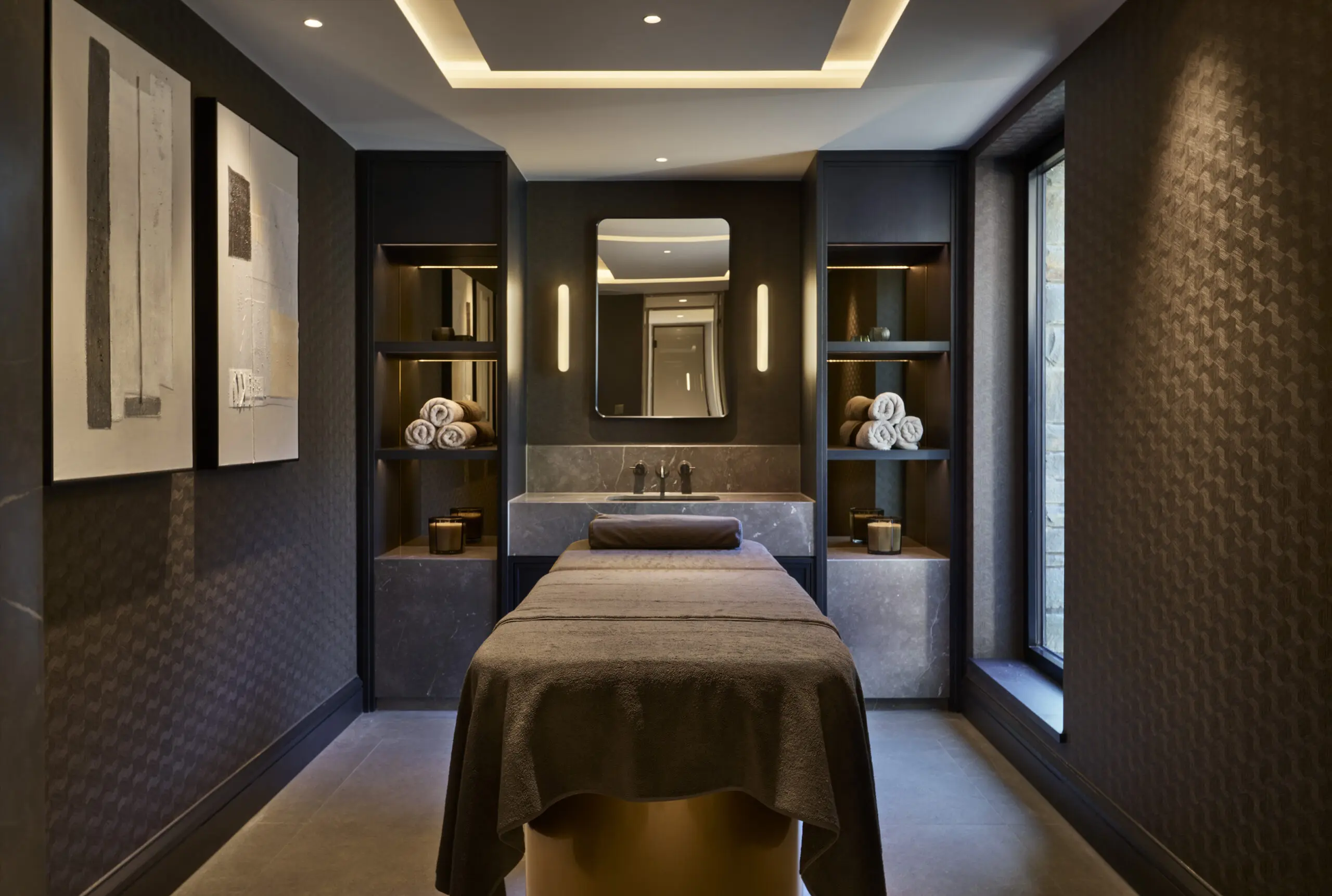 Luxurious spa room with a massage table, dim lighting, modern artwork, and neatly stacked towels. The ambiance is tranquil and inviting.