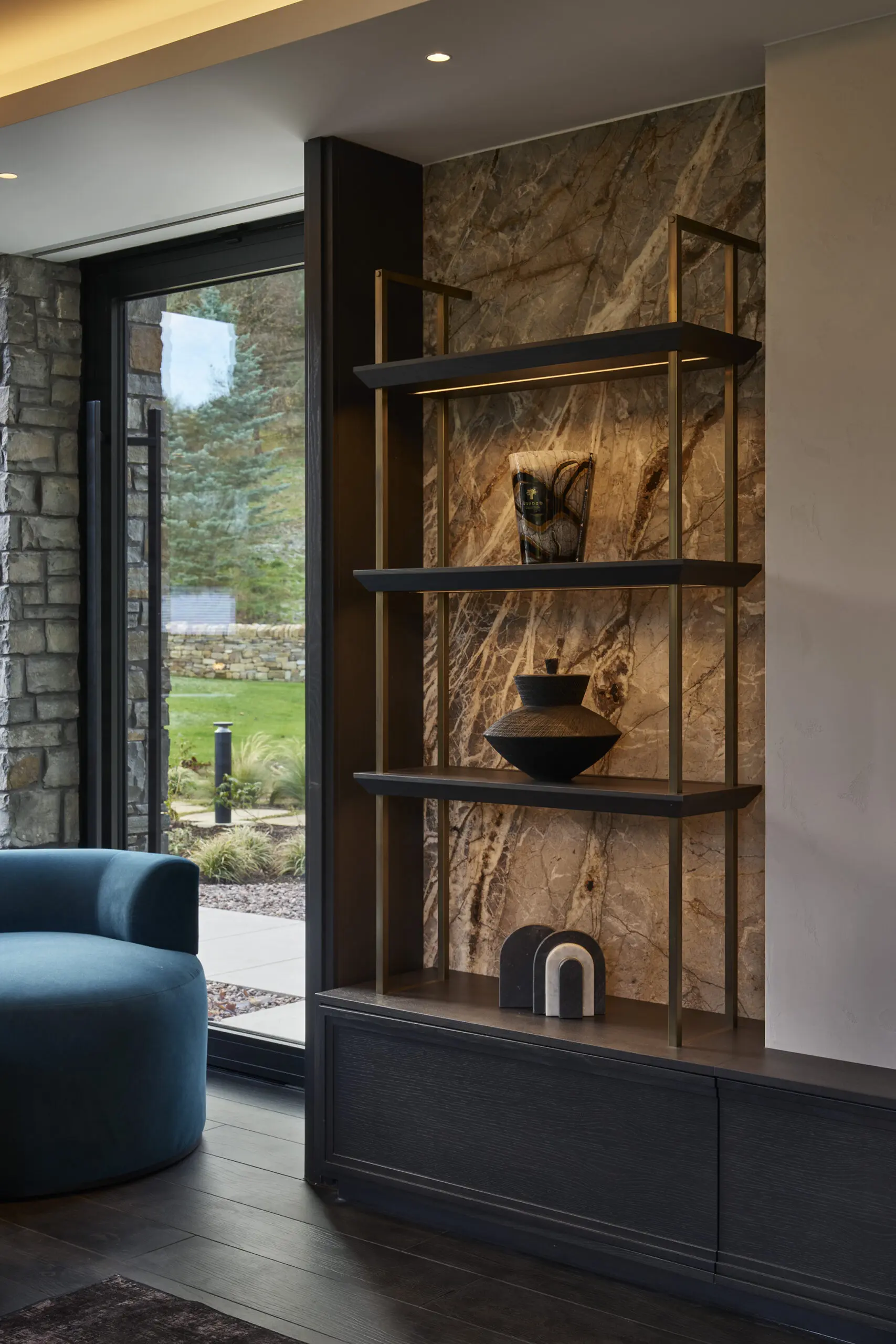 Modern interior with a stone-backed shelving unit holding vases. A large window shows a garden, and a blue chair adds a touch of color.