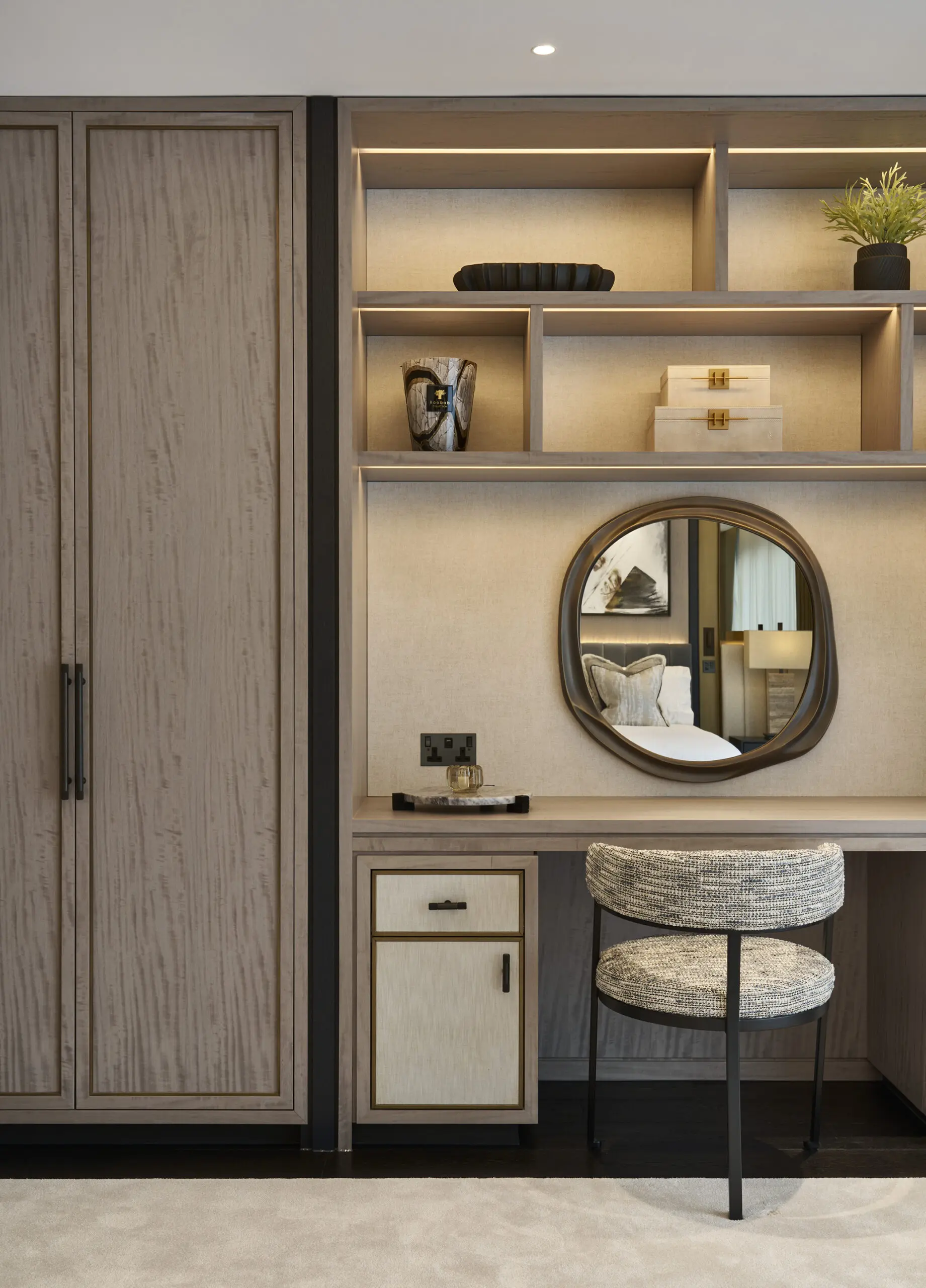 Contemporary built-in vanity and wardrobe area featuring light wood cabinetry, illuminated open shelving with decorative accents, an organic-shaped wall mirror, and a textured upholstered chair in a warm neutral-toned interior