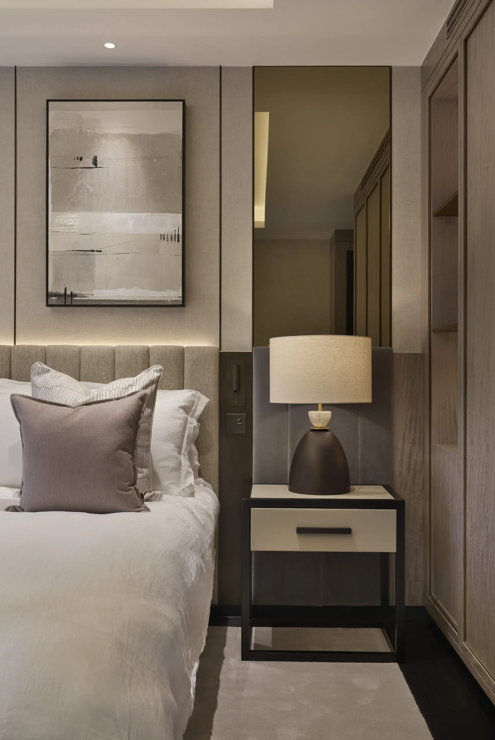 Modern neutral-toned bedroom featuring a neatly made bed with layered pillows, a minimalist bedside table with a dark ceramic lamp, vertical wall paneling, abstract framed artwork, and warm ambient lighting beside a built-in wood wardrobe